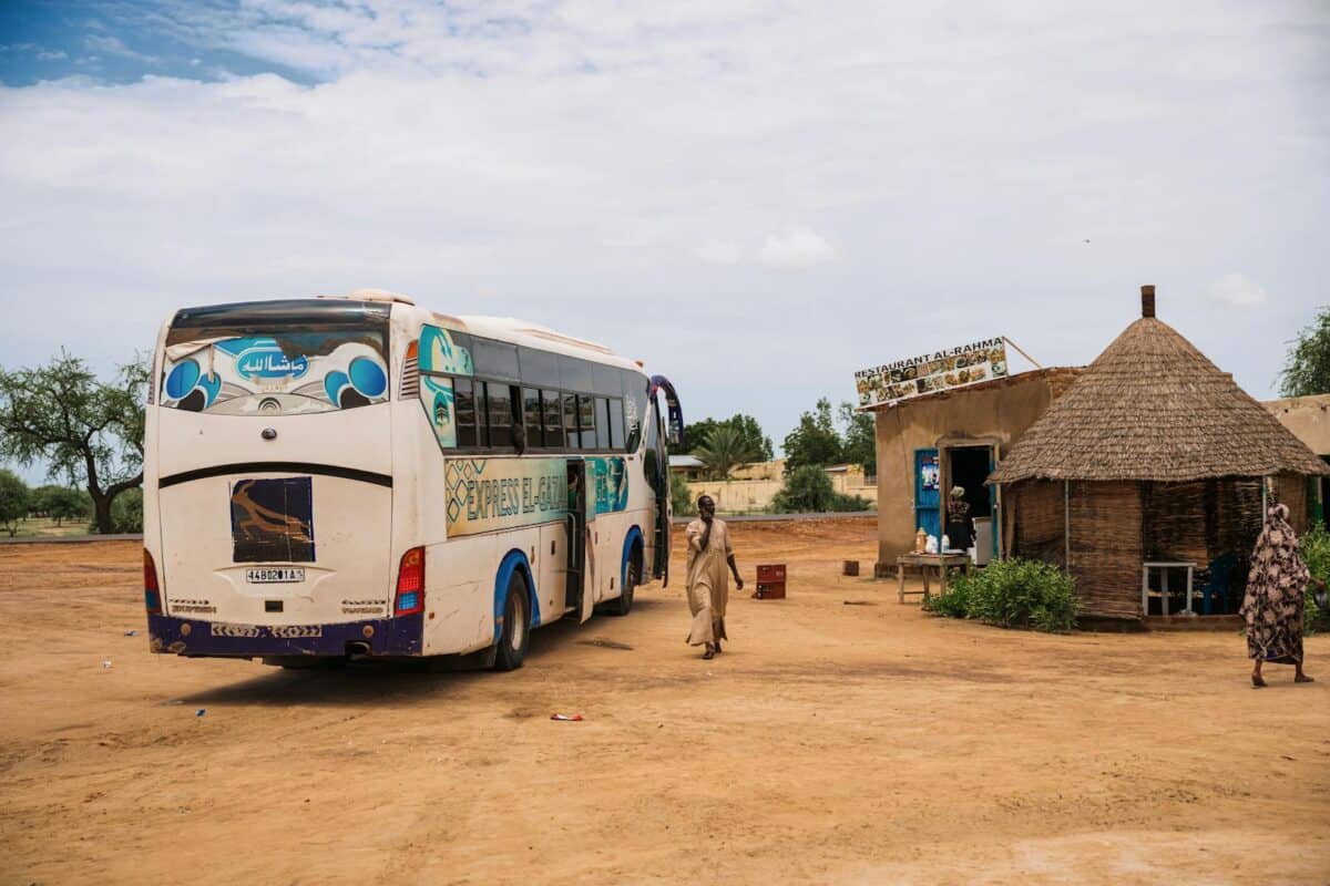 A white bus stops at a rural village with a tribal hut and local restaurant.