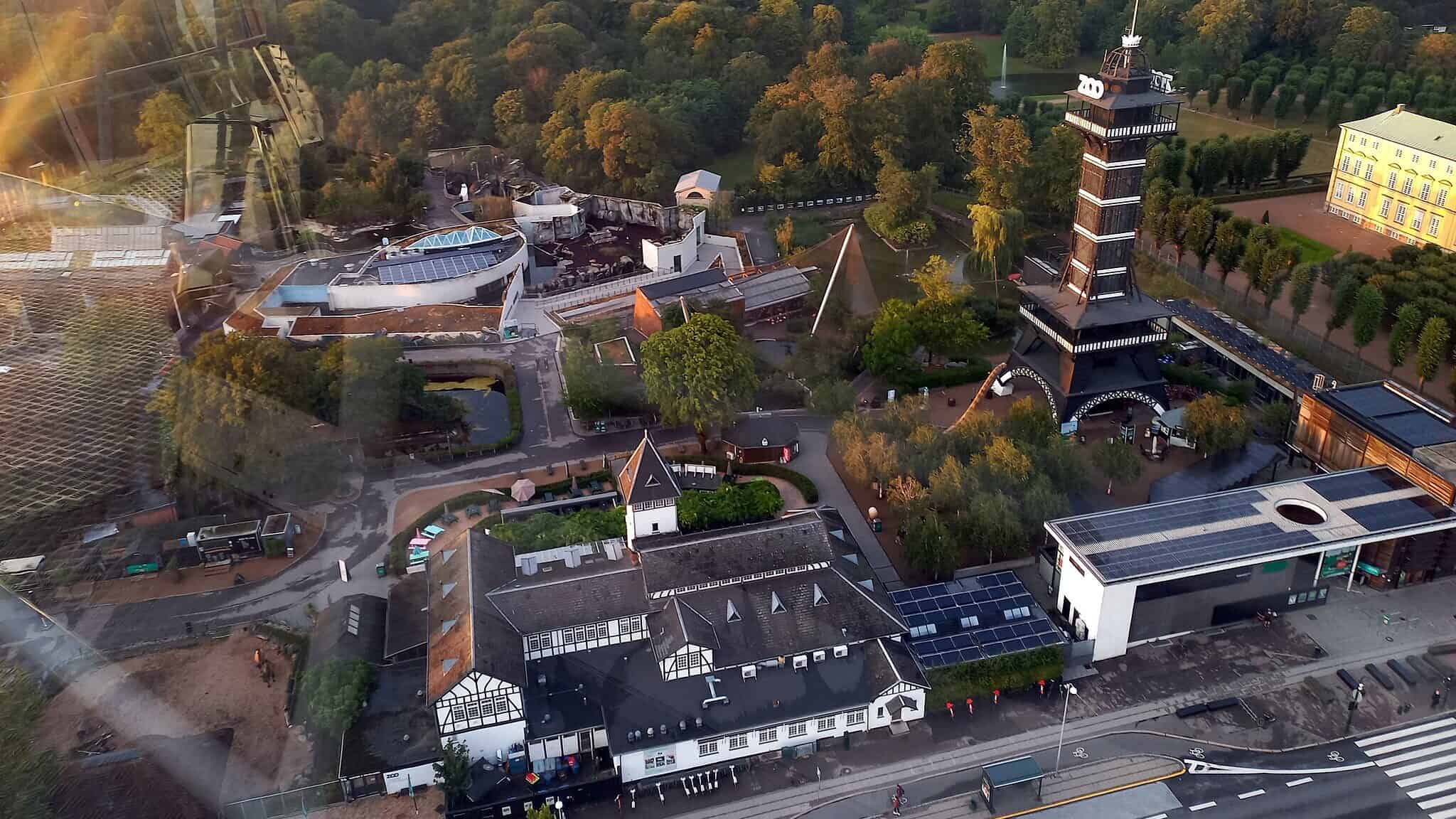 Aerial view of Copenhagen Zoo, showing its layout with lush green spaces, modern buildings, and the distinctive Zoo Tower. The grounds feature enclosures, pathways, and surrounding greenery, with the cityscape visible in the background.