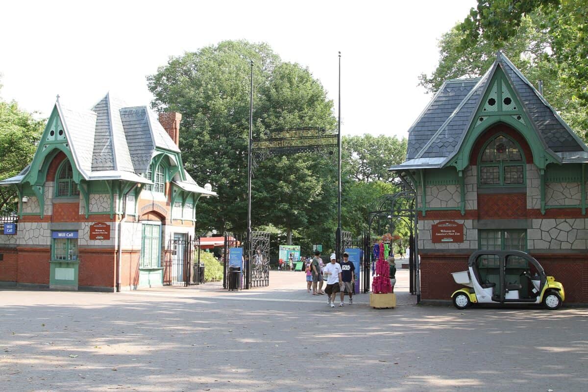 The entrance to the Philadelphia Zoo, featuring two charming, Victorian-style buildings with green and red accents on either side of a pathway. People are walking towards the entrance under a decorative wrought-iron archway. Trees and greenery are visible in the background.