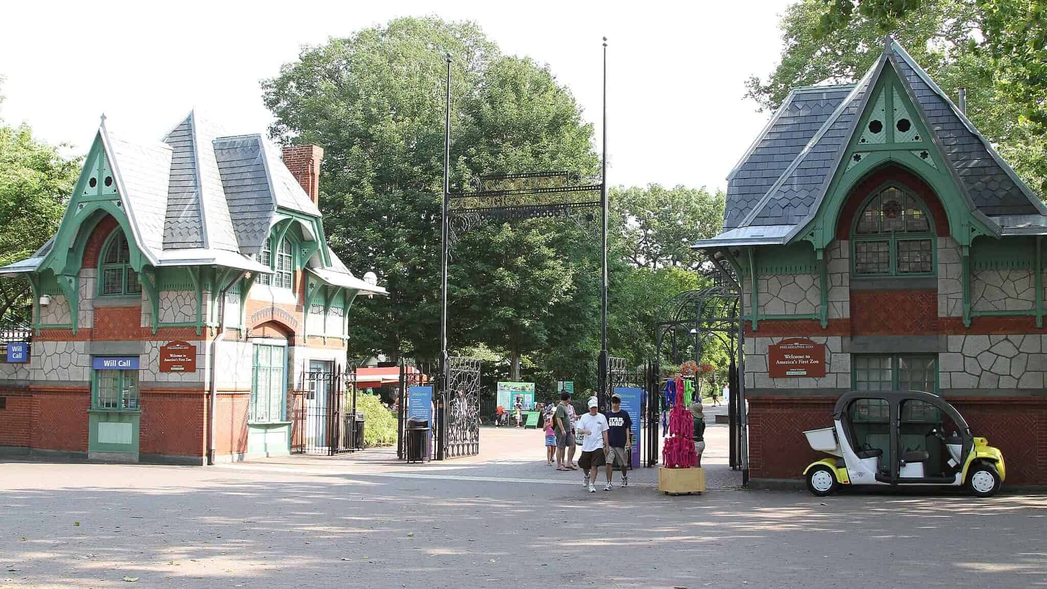 The entrance to the Philadelphia Zoo, featuring two charming, Victorian-style buildings with green and red accents on either side of a pathway. People are walking towards the entrance under a decorative wrought-iron archway. Trees and greenery are visible in the background.