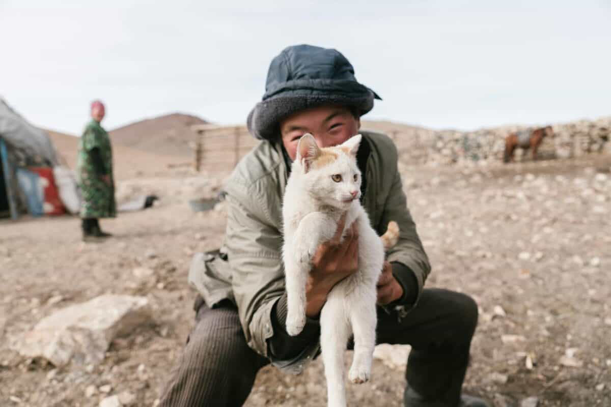 Smiling teen holding a kitten in a Mongolian desert with yurts in the background, capturing a joyful lifestyle moment.