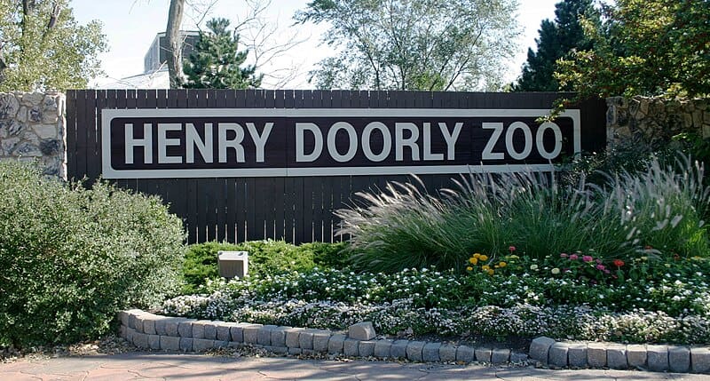 Entrance sign of the Henry Doorly Zoo, displayed on a wooden fence with lush greenery and flowers in the foreground.