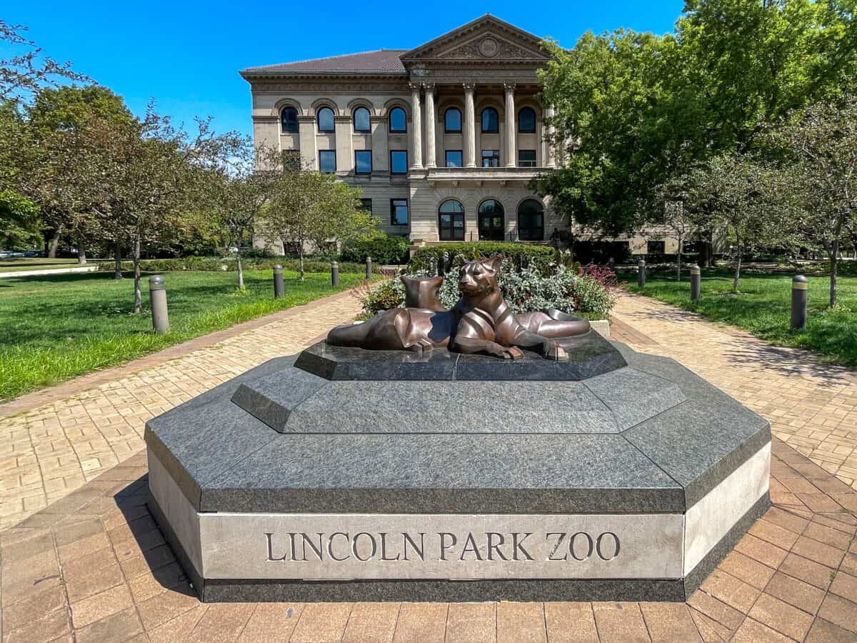A statue of two reclining lions on a granite base inscribed with "Lincoln Park Zoo," surrounded by a landscaped pathway with greenery and trees in the background.
