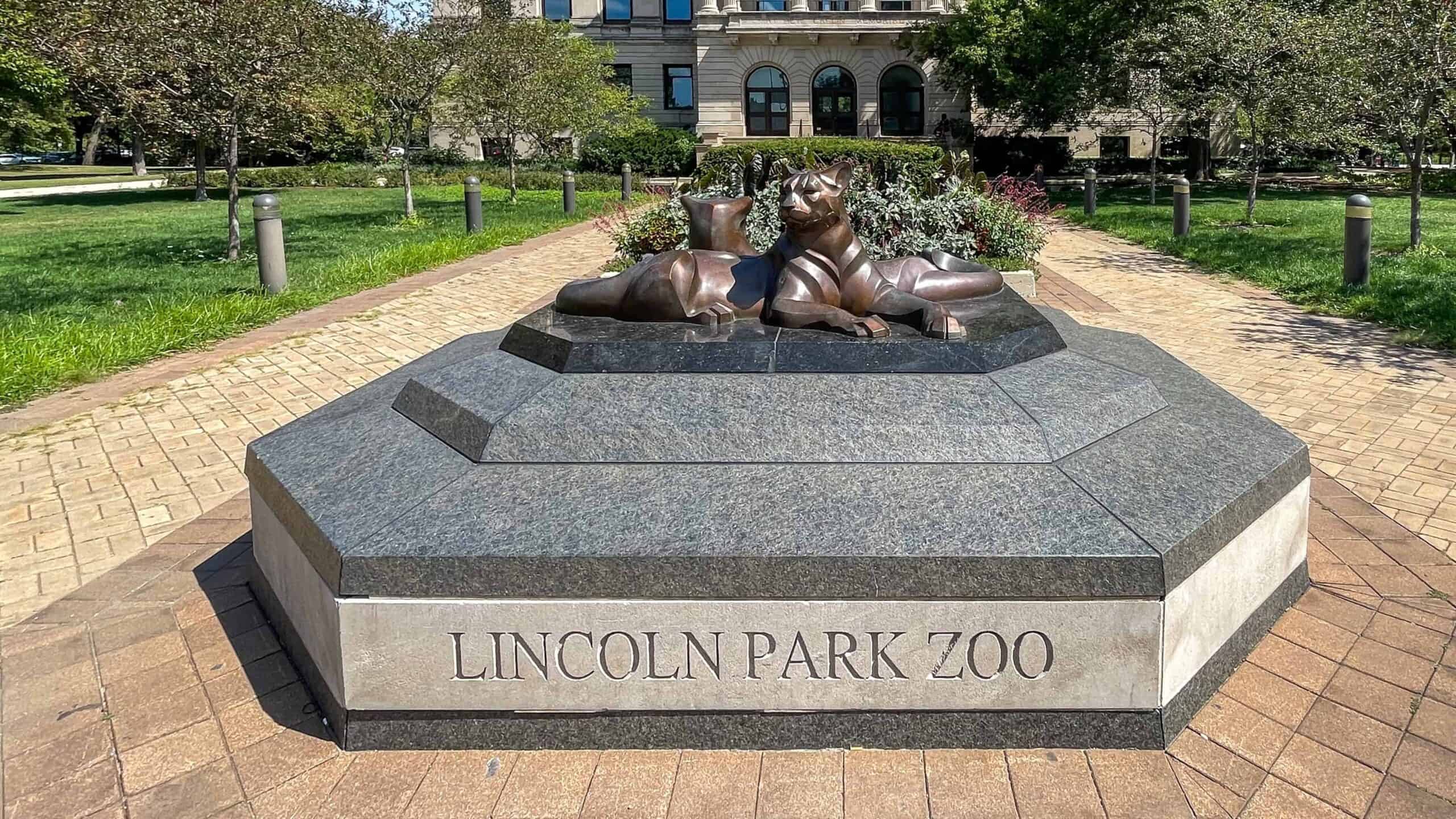 A statue of two reclining lions on a granite base inscribed with "Lincoln Park Zoo," surrounded by a landscaped pathway with greenery and trees in the background.