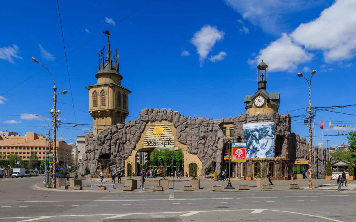 A grand entrance to a zoo or theme park, featuring castle-like towers, stone-like structures, and decorative elements under a bright blue sky.