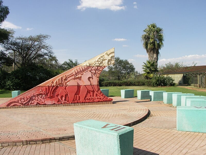 A sundial monument at the National Zoological Gardens of South Africa in Pretoria, featuring animal carvings and surrounded by numbered turquoise seating blocks.