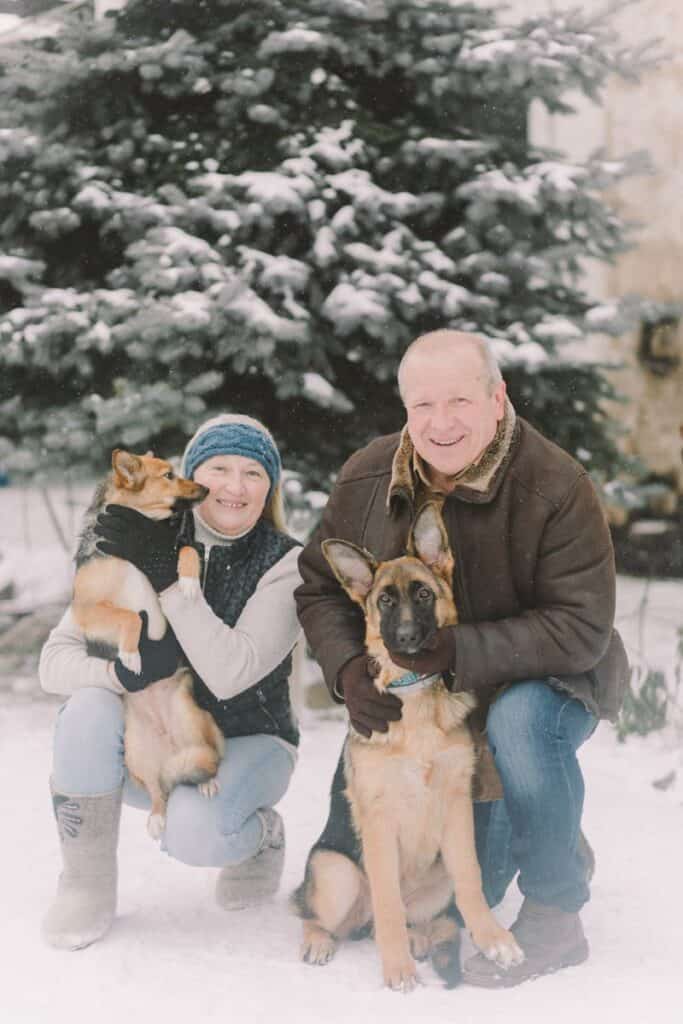 Senior couple with German Shepherd and another dog outdoors in snowy winter setting.