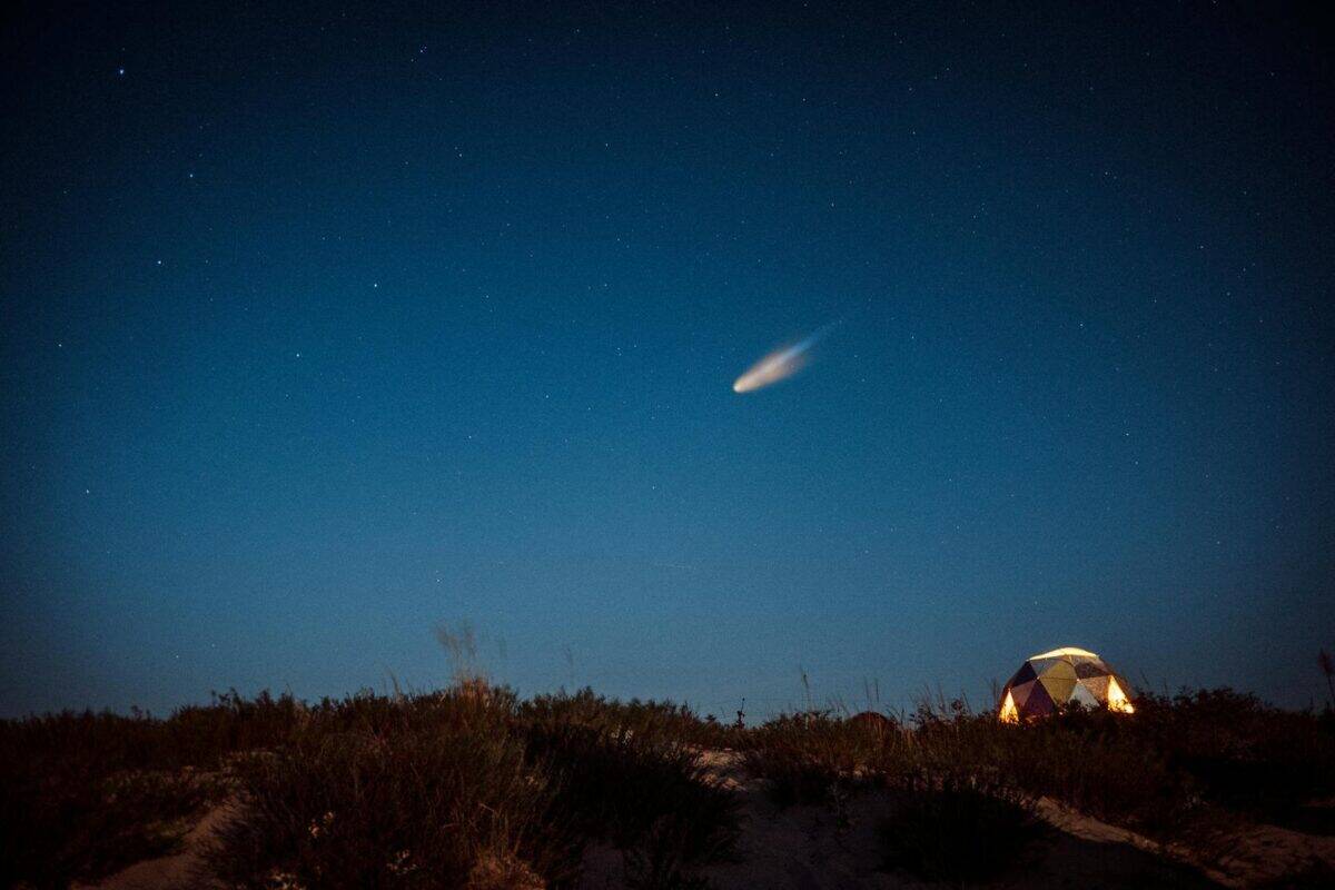 Stunning night scene of a meteorite illuminating the starry sky above an illuminated tent in a serene landscape.