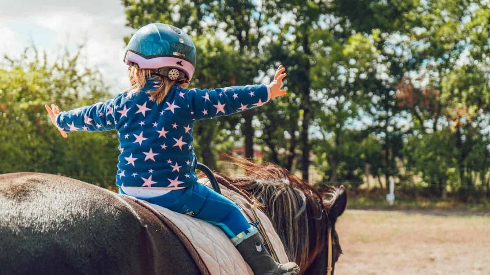 A child confidently rides a horse outdoors, wearing a helmet and star-patterned jacket.