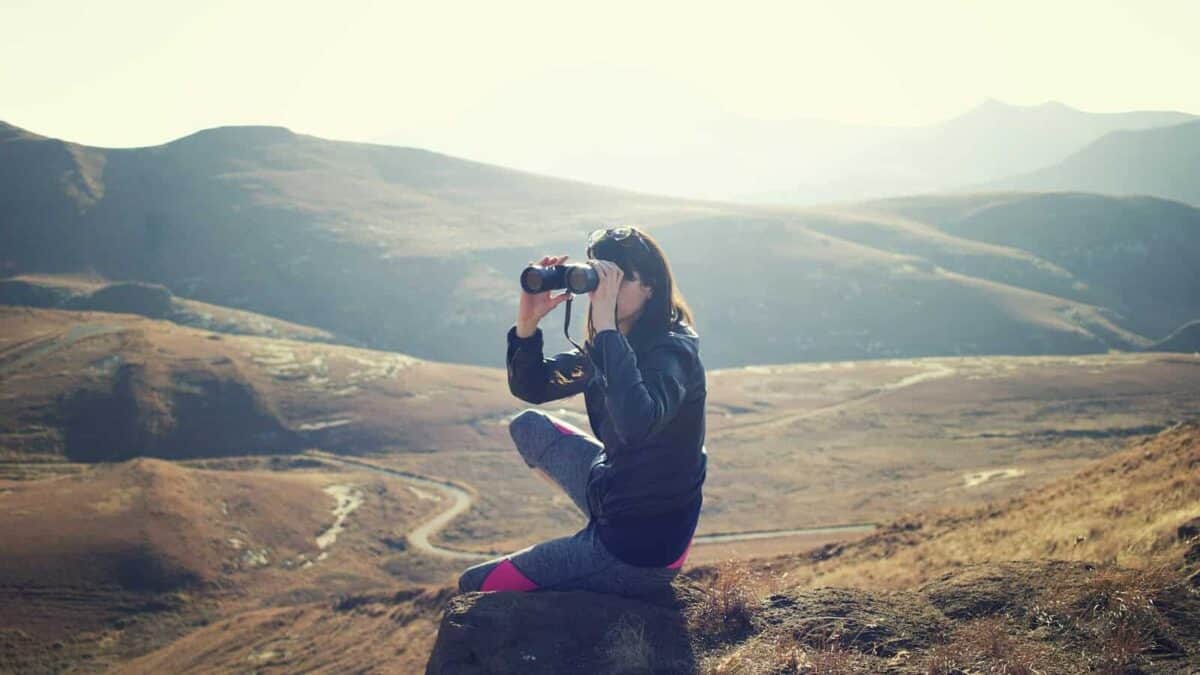 woman wearing black jacket sitting on gray rock and using binoculrs