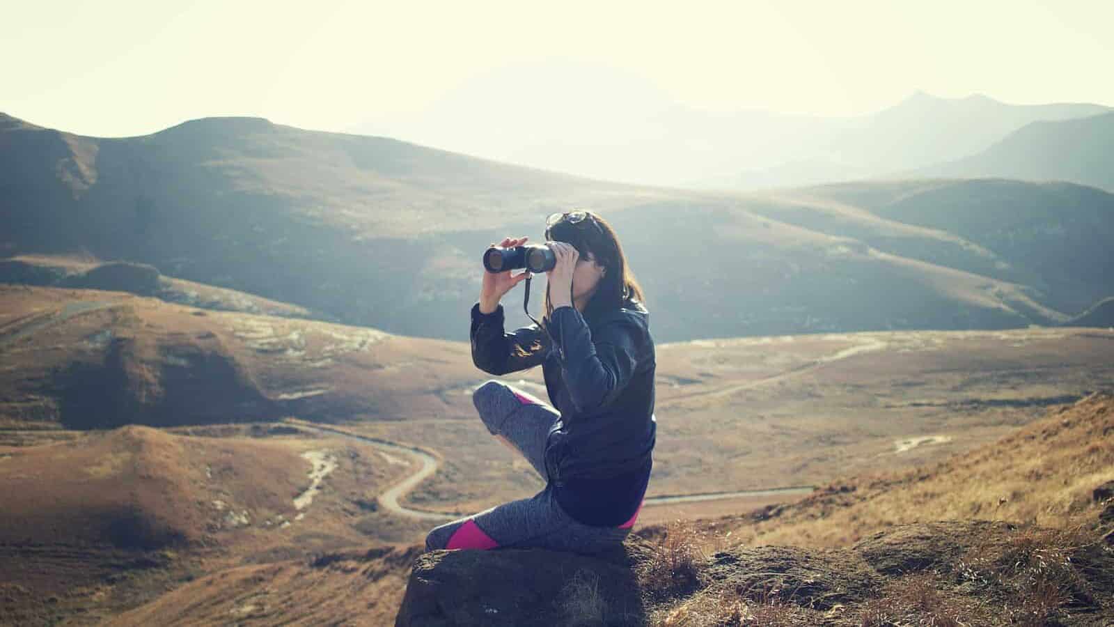 woman wearing black jacket sitting on gray rock and using binoculrs