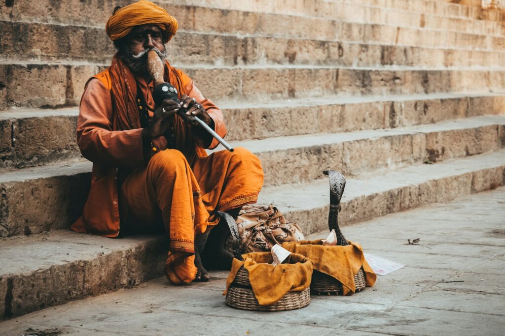 A man in an orange thobe sitting on concrete stairs playing a musical instrument to a cobra snake 