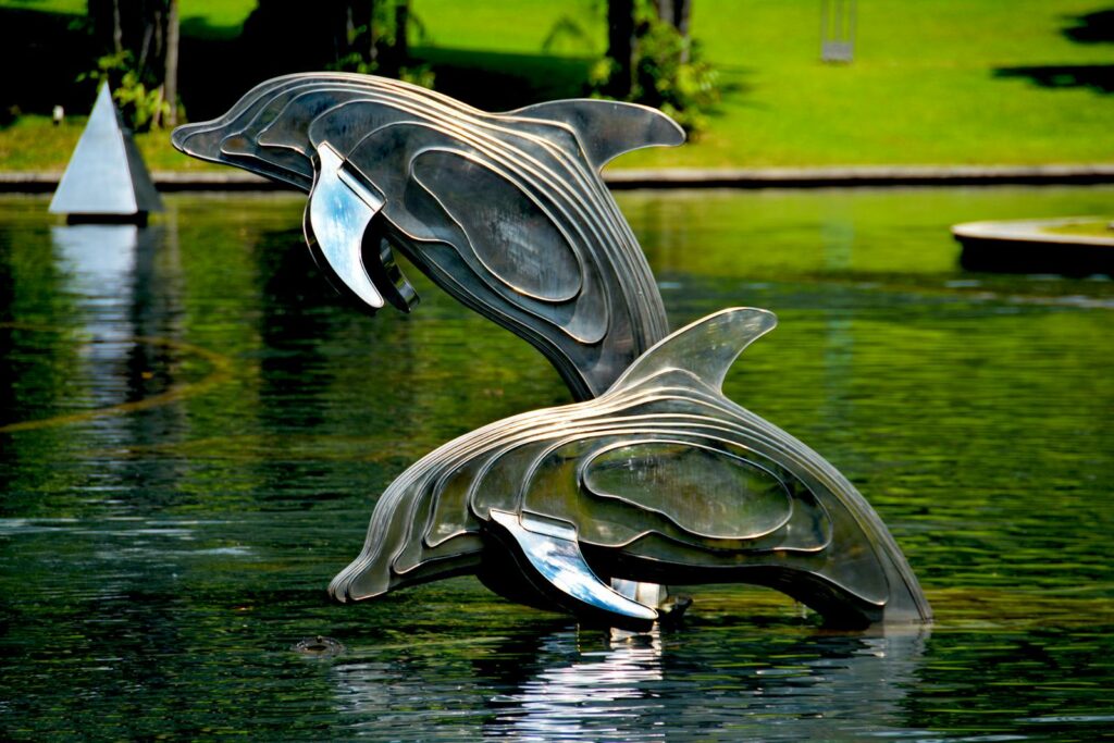 Stainless steel dolphin statues jump gracefully in a serene park pond, surrounded by greenery.