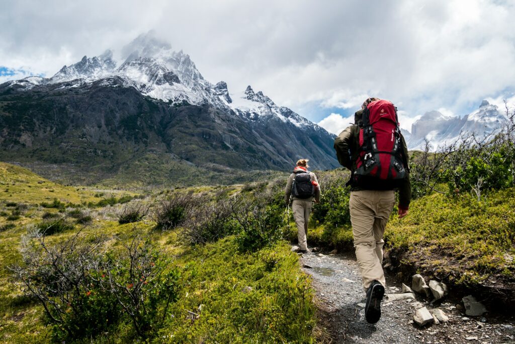 Two hikers with backpacks walking along a scenic mountain trail surrounded by lush greenery and rocky terrain.